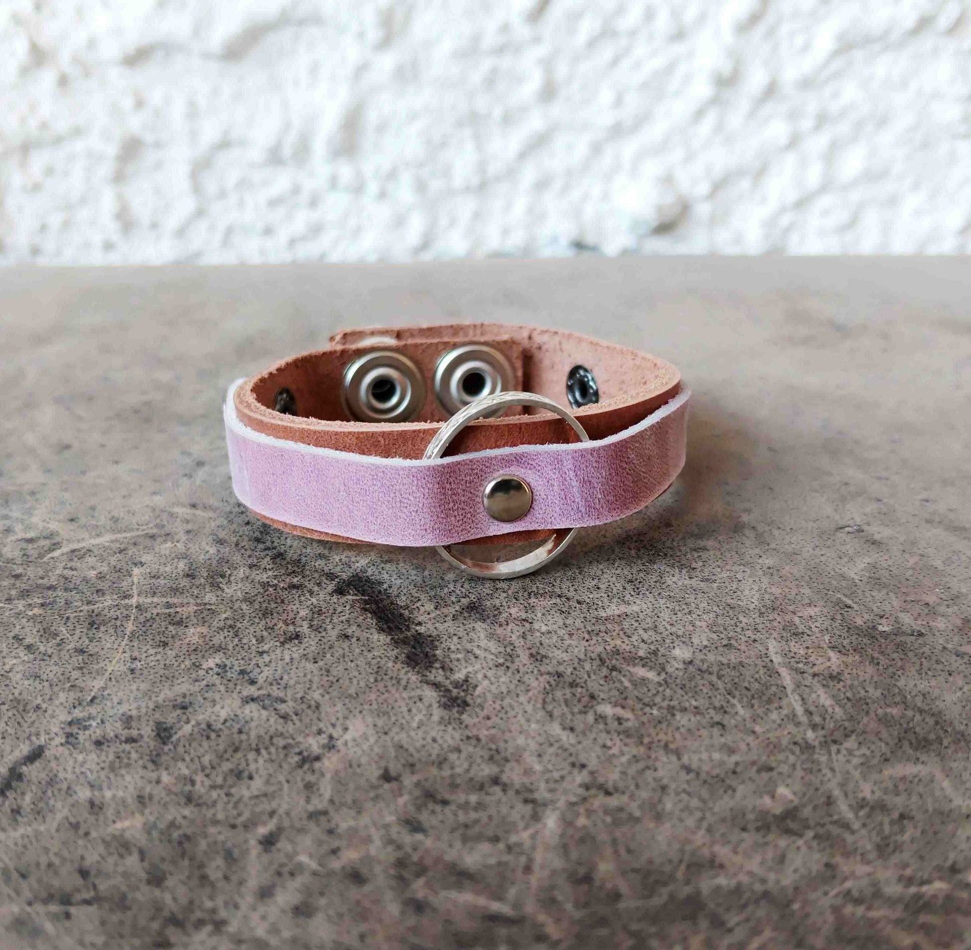 A light pink leather bracelet with a silver metal O-ring and snap button, resting on a textured gray stone surface against a white stucco wall.