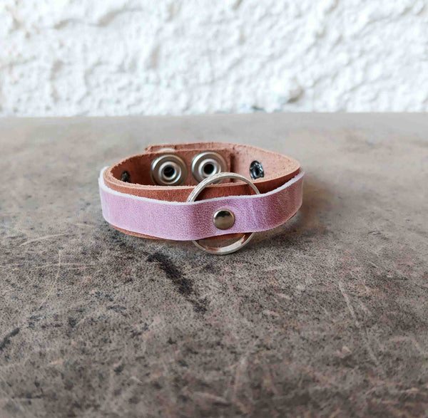 A light pink leather bracelet with a silver metal O-ring and snap button, resting on a textured gray stone surface against a white stucco wall.