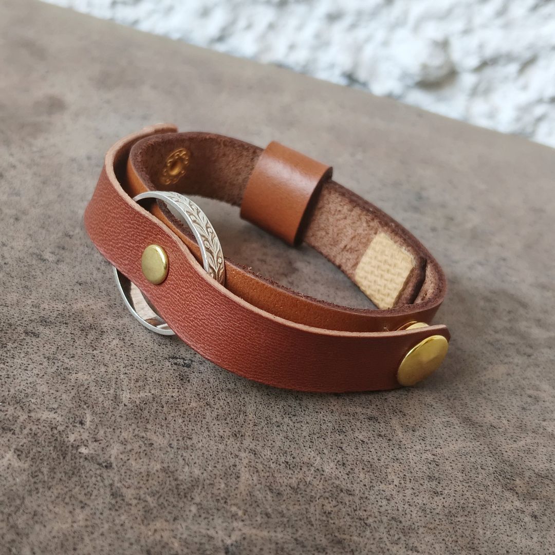 A brown leather ring holder bracelet with brass and silver-tone metal accents, displayed on a textured gray surface.