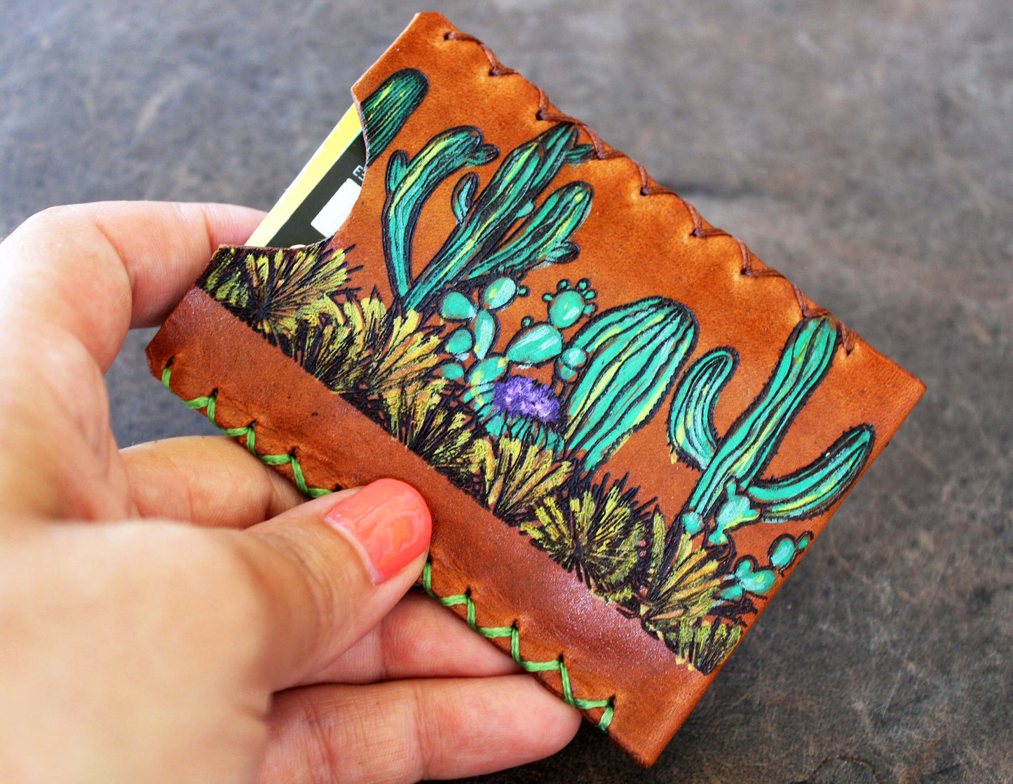 Hand holding a leather cardholder with cactus design on a dark surface