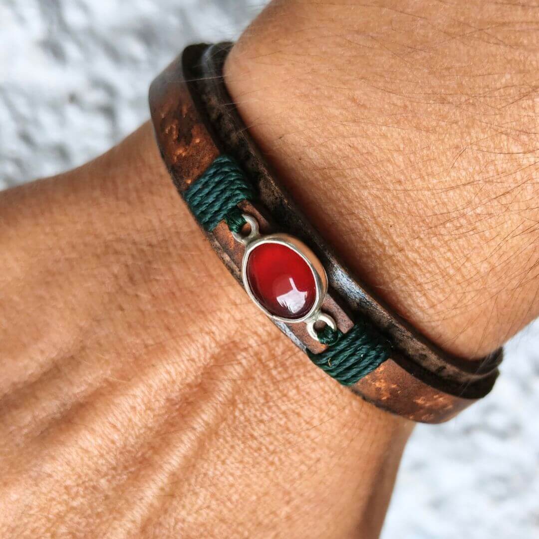 A close-up of a person's wrist wearing a multi-strand bracelet featuring dark brown leather, dark green cord, and a central oval red agate stone in a silver-toned setting.