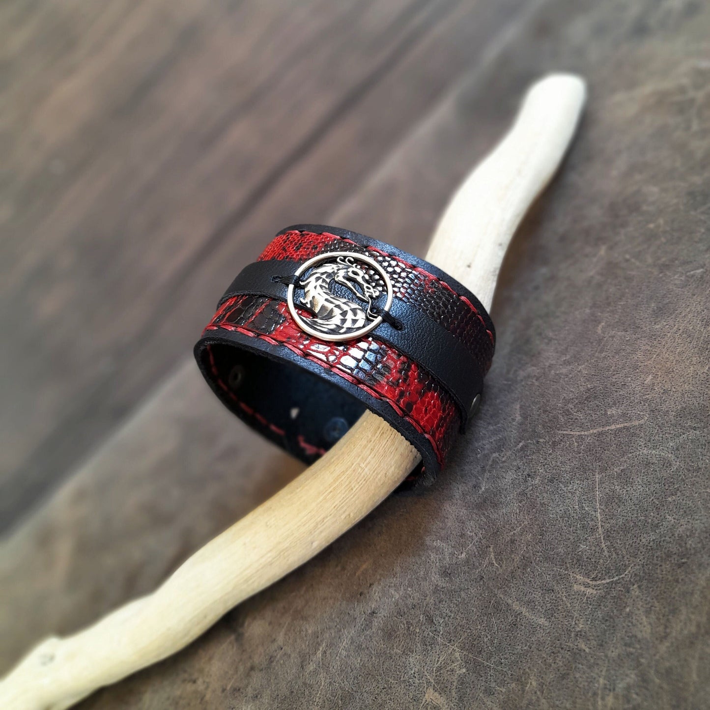 A black and red wide leather cuff with a snake print design, featuring a dragon pendant, displayed on a wooden branch.