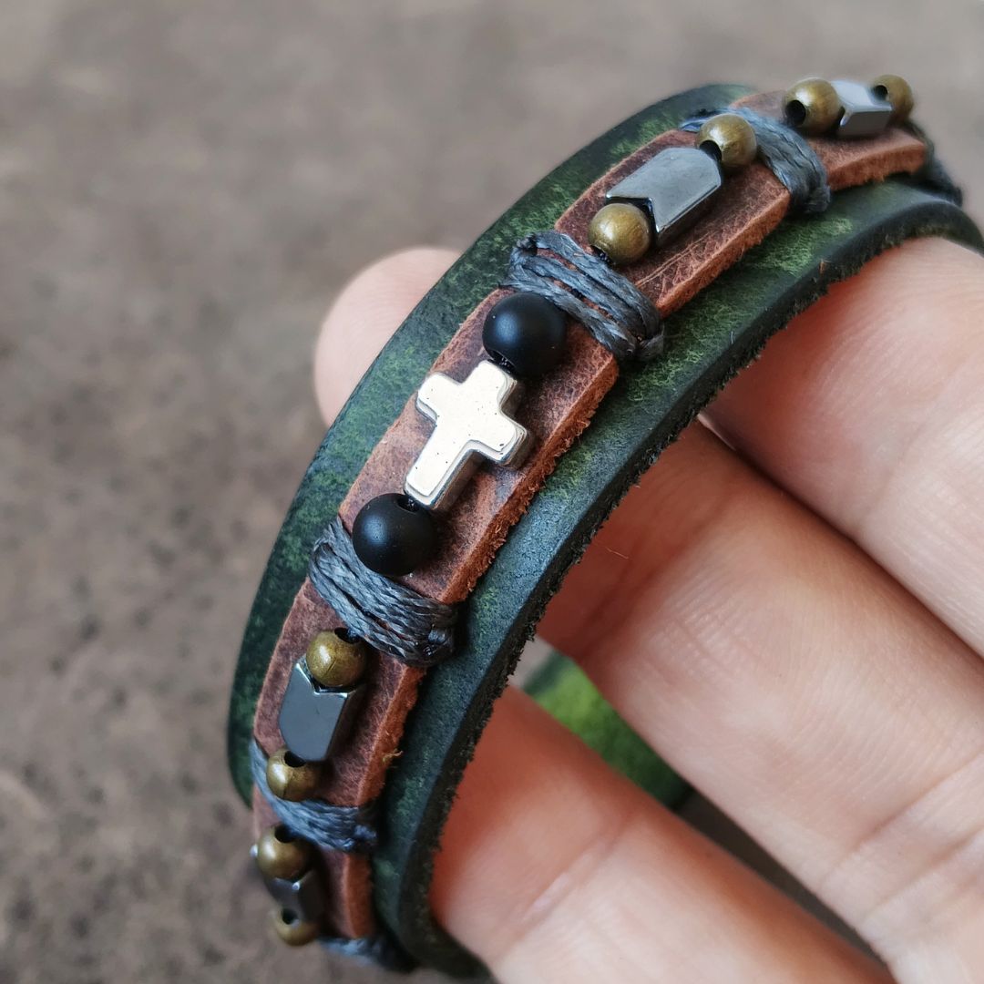 A close-up of a hand holding a multi-layered leather cuff bracelet. The bracelet features dark green and brown leather bands, wrapped with gray cord, adorned with a silver cross, black beads, and small metallic accents.