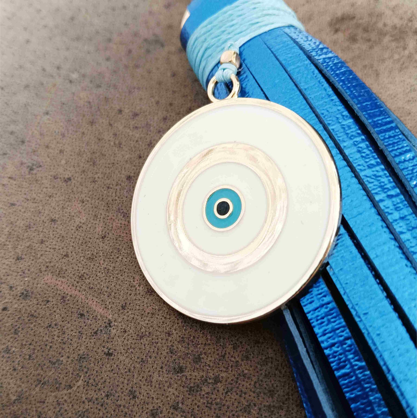 A circular white and turquoise evil eye pendant with a gold-tone border, resting on a textured brown surface next to a bright blue tassel.
