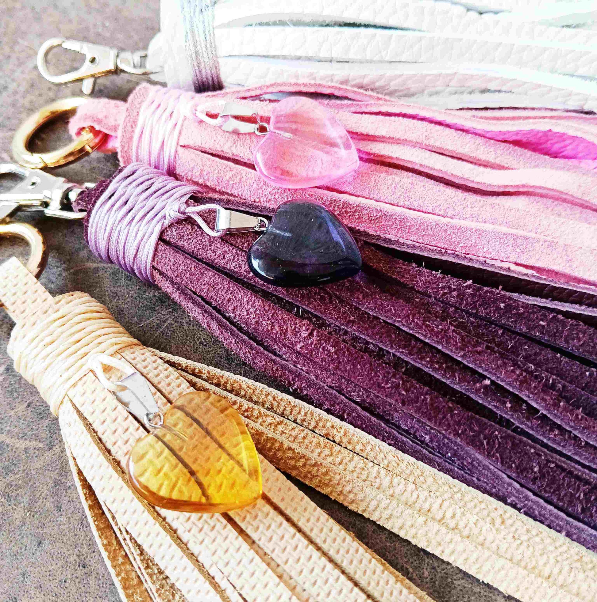 A close-up of multiple leather tassel keychains or bag charms arranged diagonally. The tassels are in cream, light pink, lavender, and deep purple, each attached to a silver clasp and decorated with a small heart-shaped charm, including one amber glass heart and one dark purple heart.