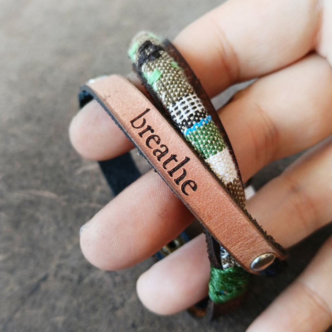 A close-up of a hand holding a multi-layered leather cuff bracelet. The bracelet features dark green and brown leather bands.