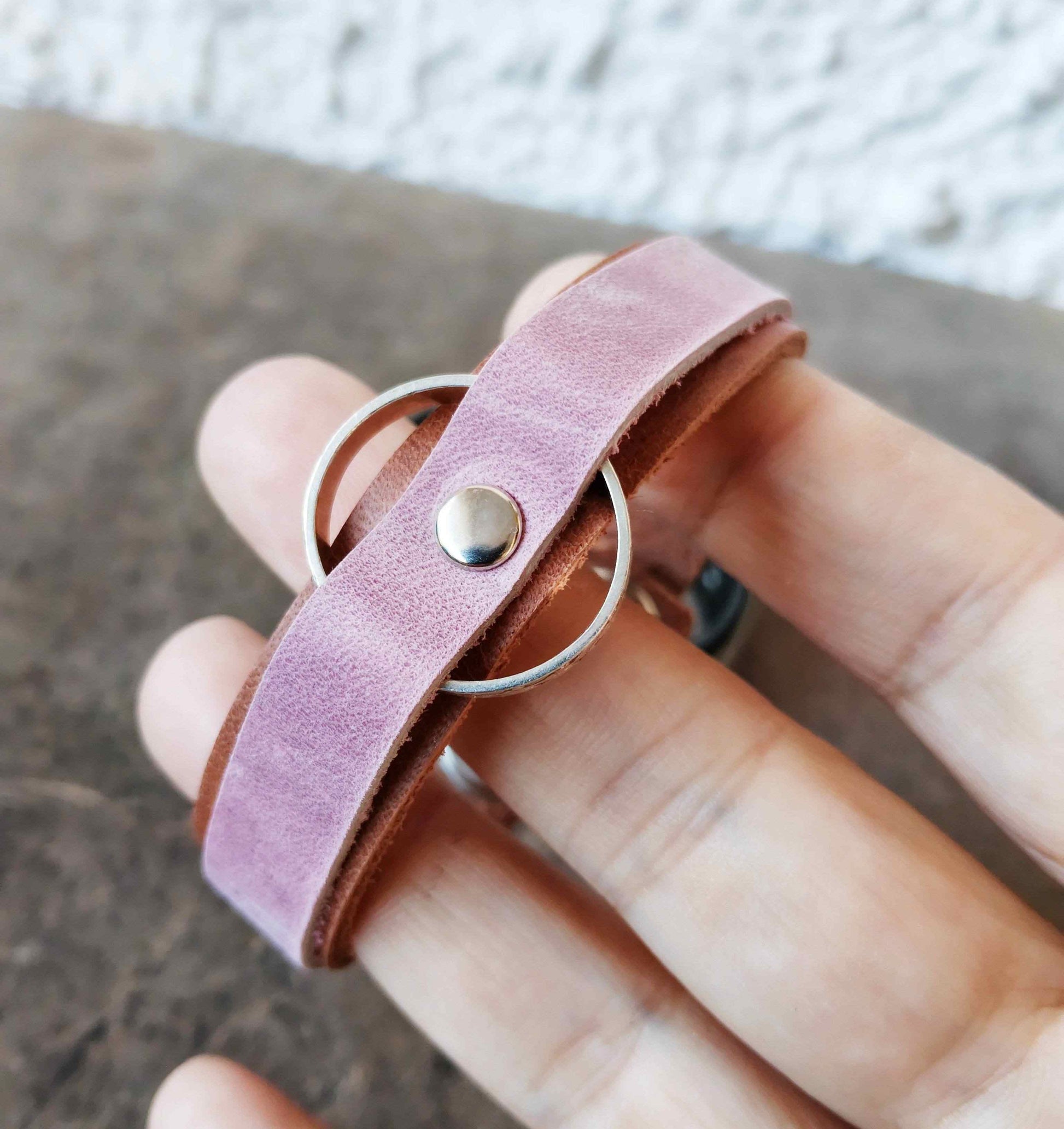 A close-up of a hand holding a pink leather wrap bracelet with a silver metal ring and a round stud.
