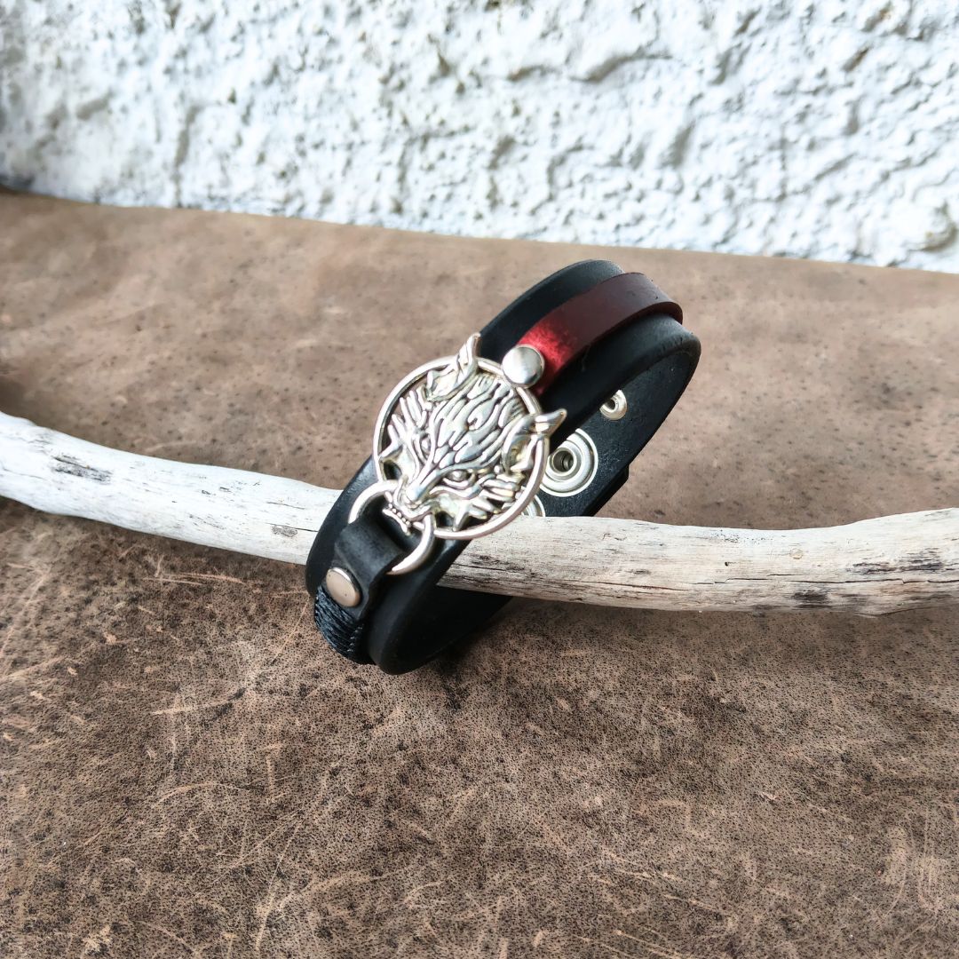 A handmade leather cuff bracelet with a central round bezel containing a red cabochon stone, accented by dark blue braided cord and silver-toned metal hardware, resting on a textured brown surface.