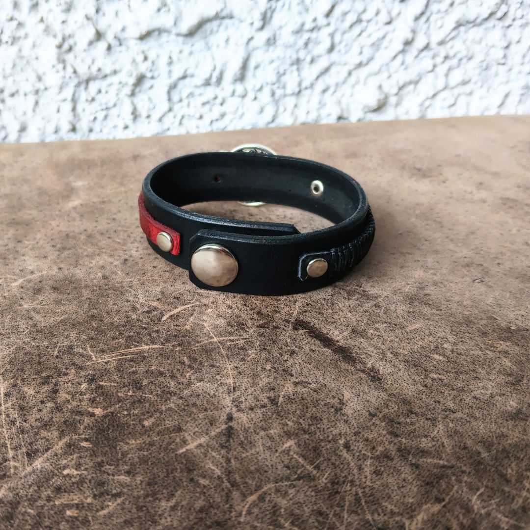 A black leather cuff bracelet with a red leather accent panel and silver-tone metal snaps, resting on a textured brown surface against a white stucco wall.