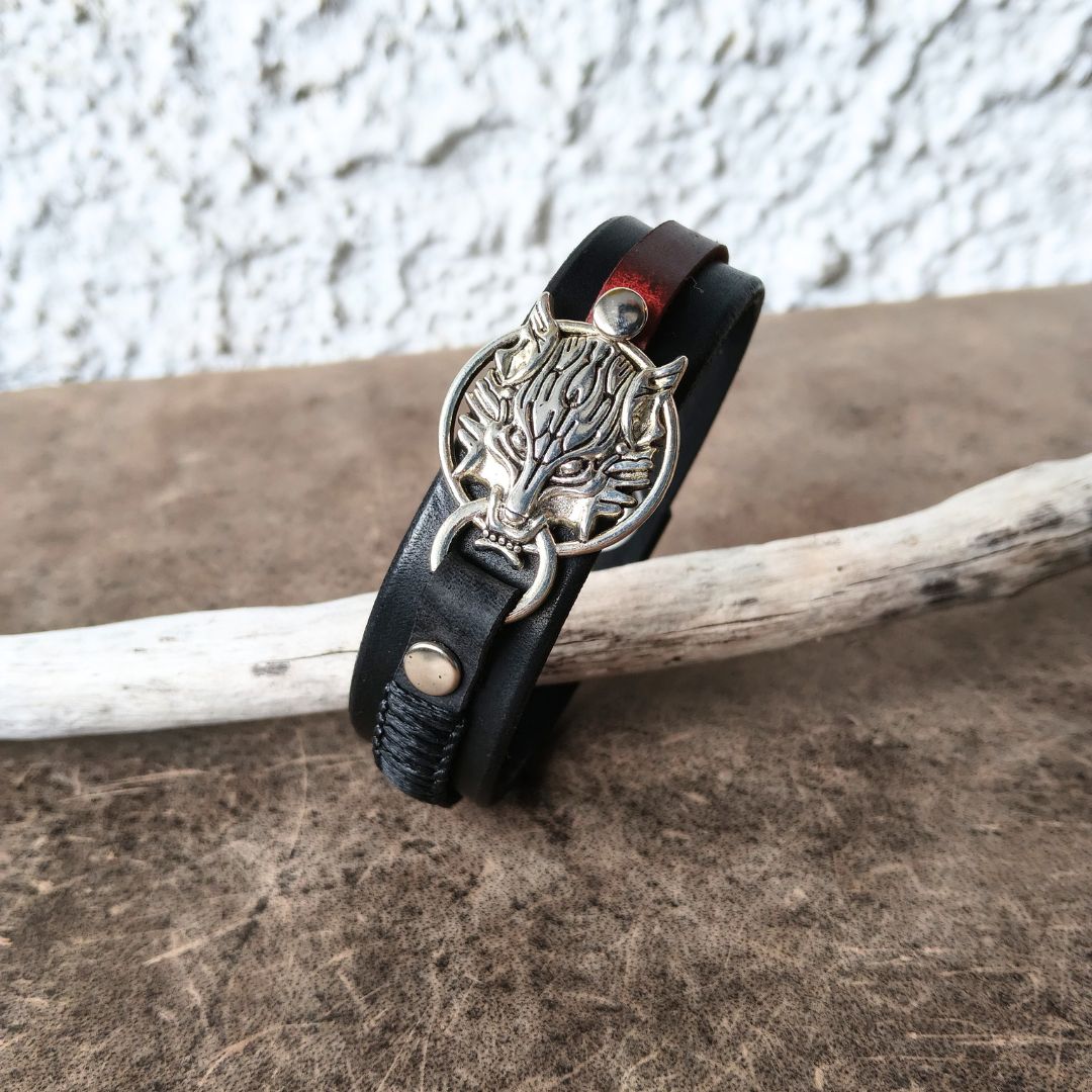 A black leather wrap bracelet with a silver-toned wolf head charm and snap closure, displayed on a rustic wooden surface with a white textured wall in the background.