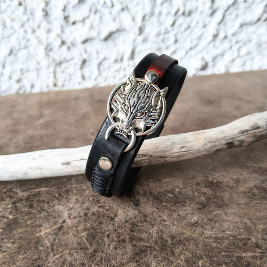 A black leather wrap bracelet with a silver-toned wolf head charm and snap closure, displayed on a rustic wooden surface with a white textured wall in the background.