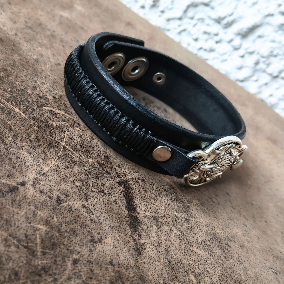 A black leather wrap bracelet with a textured braided section, silver-tone snap buttons, and a decorative silver-toned metal clasp, displayed on a textured brown surface.
