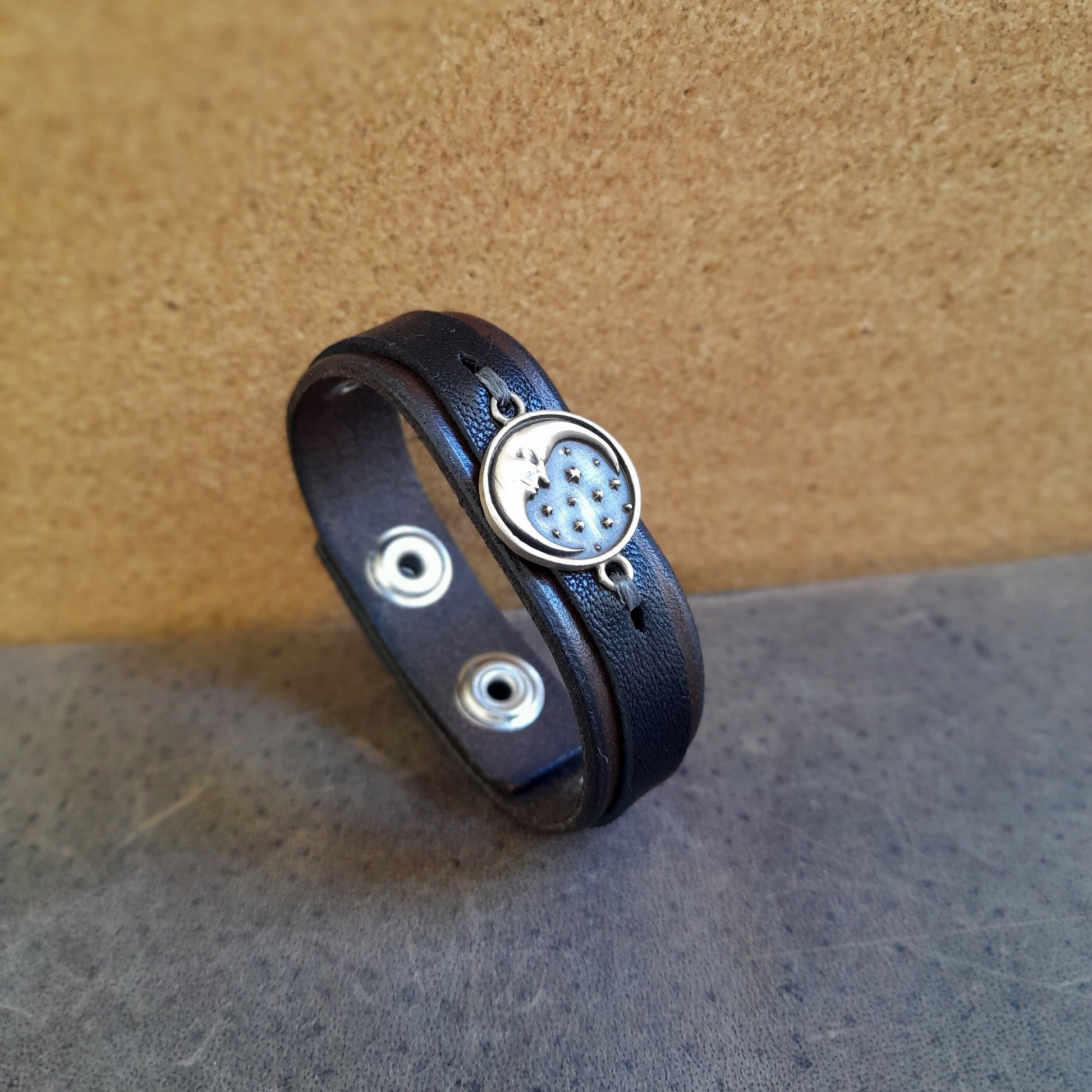 A chestnut brown and black leather bracelet with a crescent moon oxidized brass pendant, displayed on a textured background.