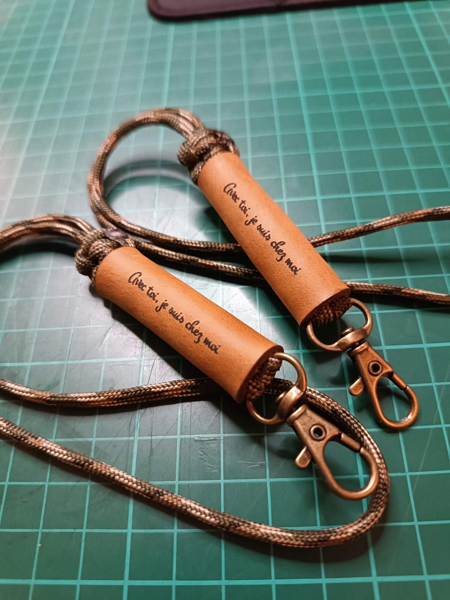 Two brown leather keychains with rope straps on a green cutting mat.