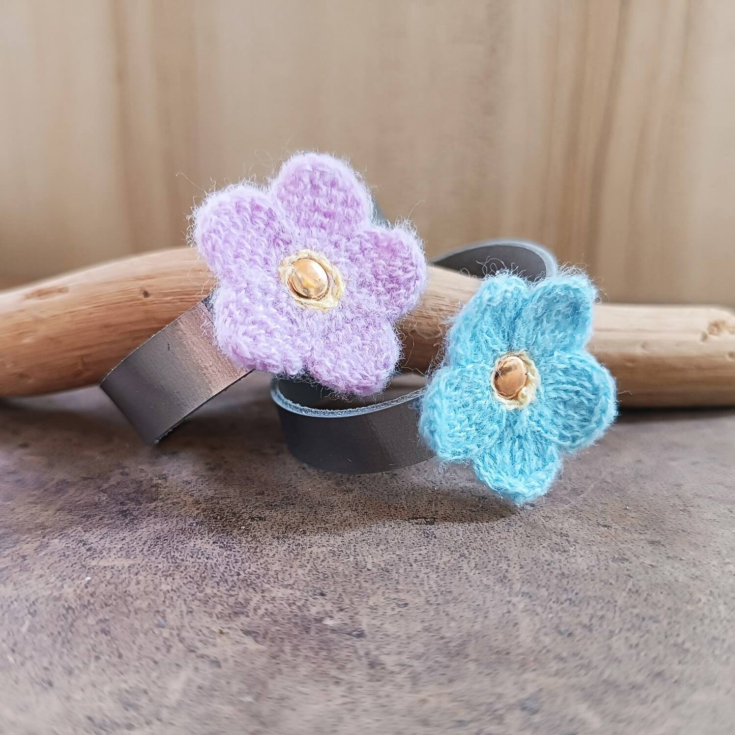 Two knitted flower-shaped on silver leather bands, one purple and one blue, on a wooden surface.