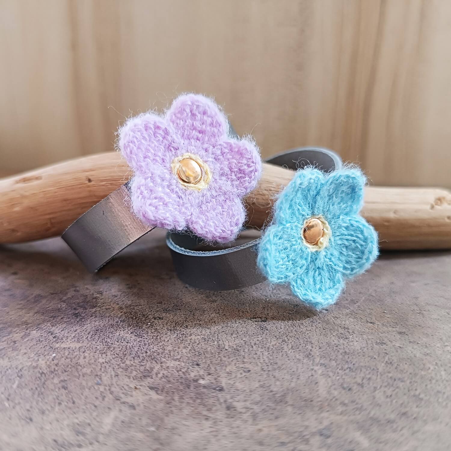 Two knitted flower-shaped on silver leather bands, one purple and one blue, on a wooden surface.