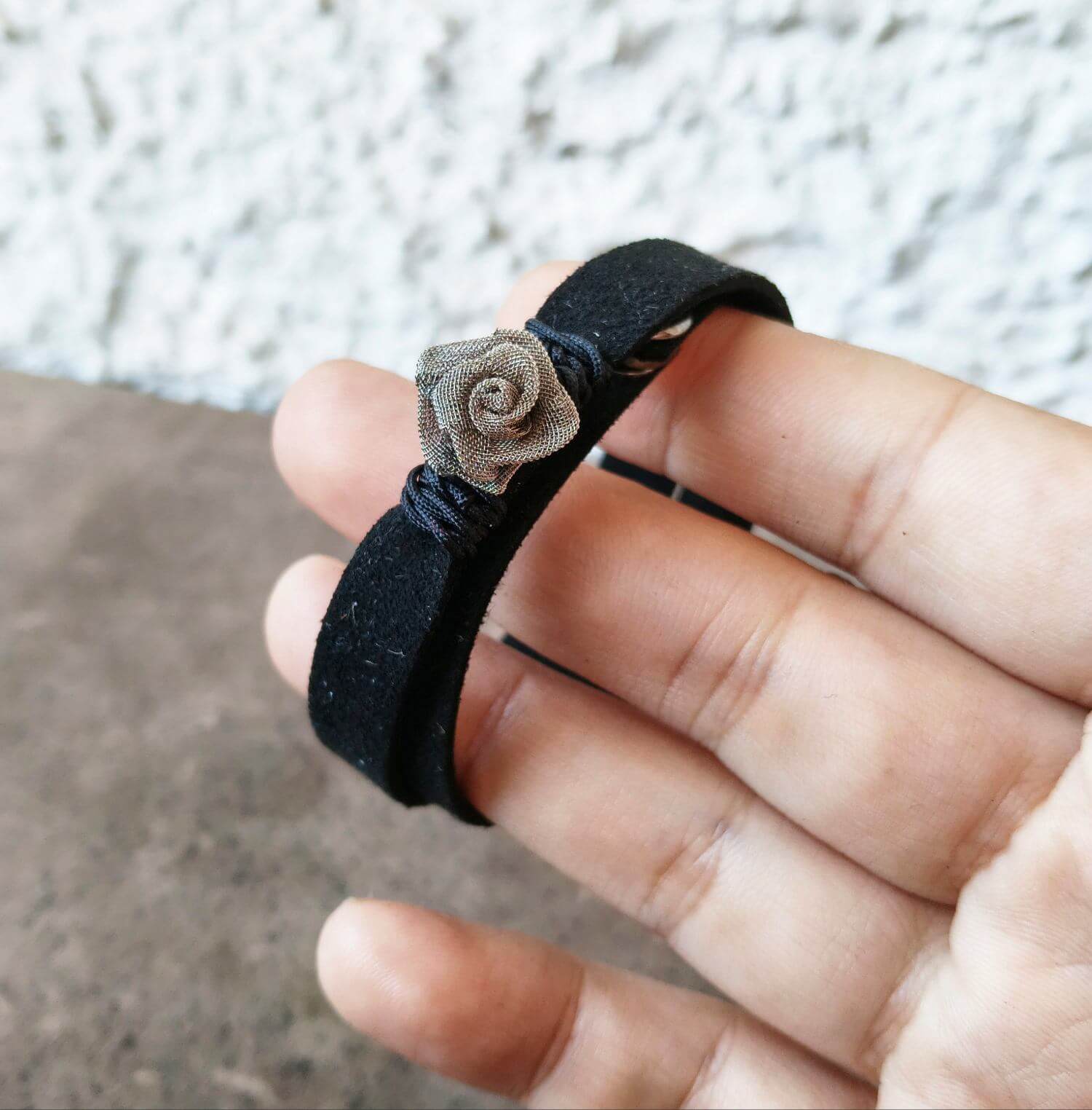 A close-up of a hand holding a black suede double wrap bracelet with a small wire rose embellishment, set against a blurred outdoor background.