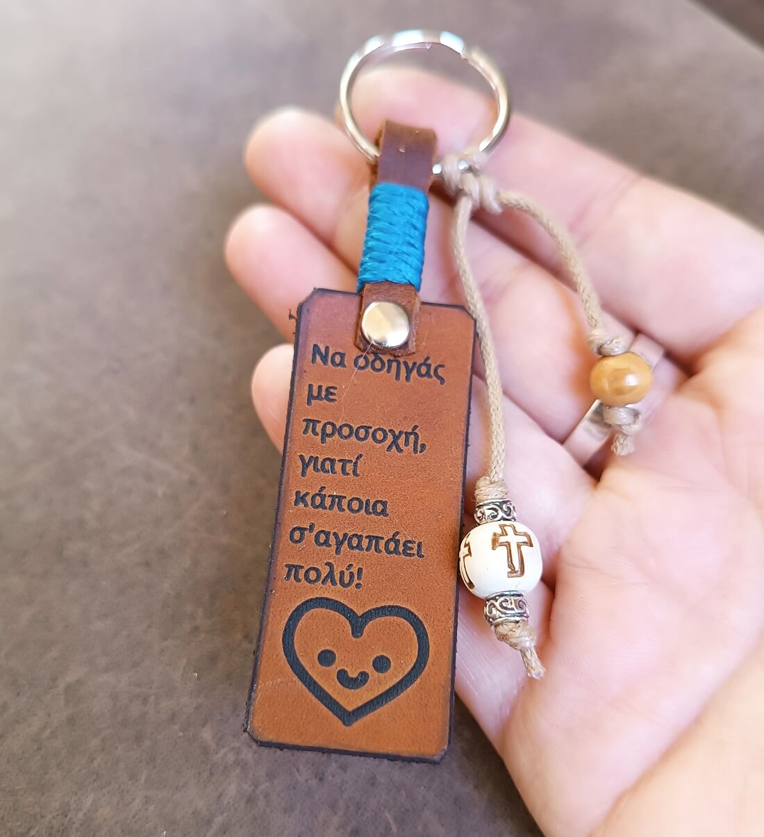 Brown leather keychain with Greek text and heart design held by a hand against a neutral background
