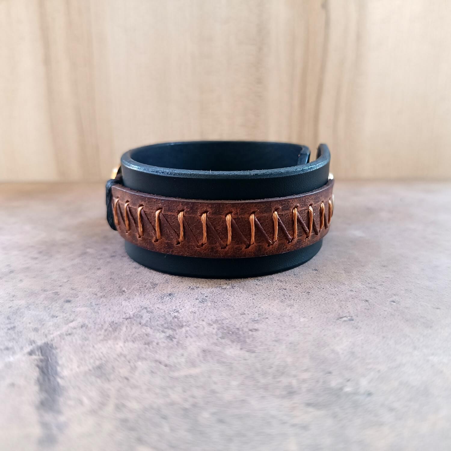 Side view of wide black and brown leather cuff bracelet, showing thick leather layers, detailed zigzag stitching, and adjustable snap closure.