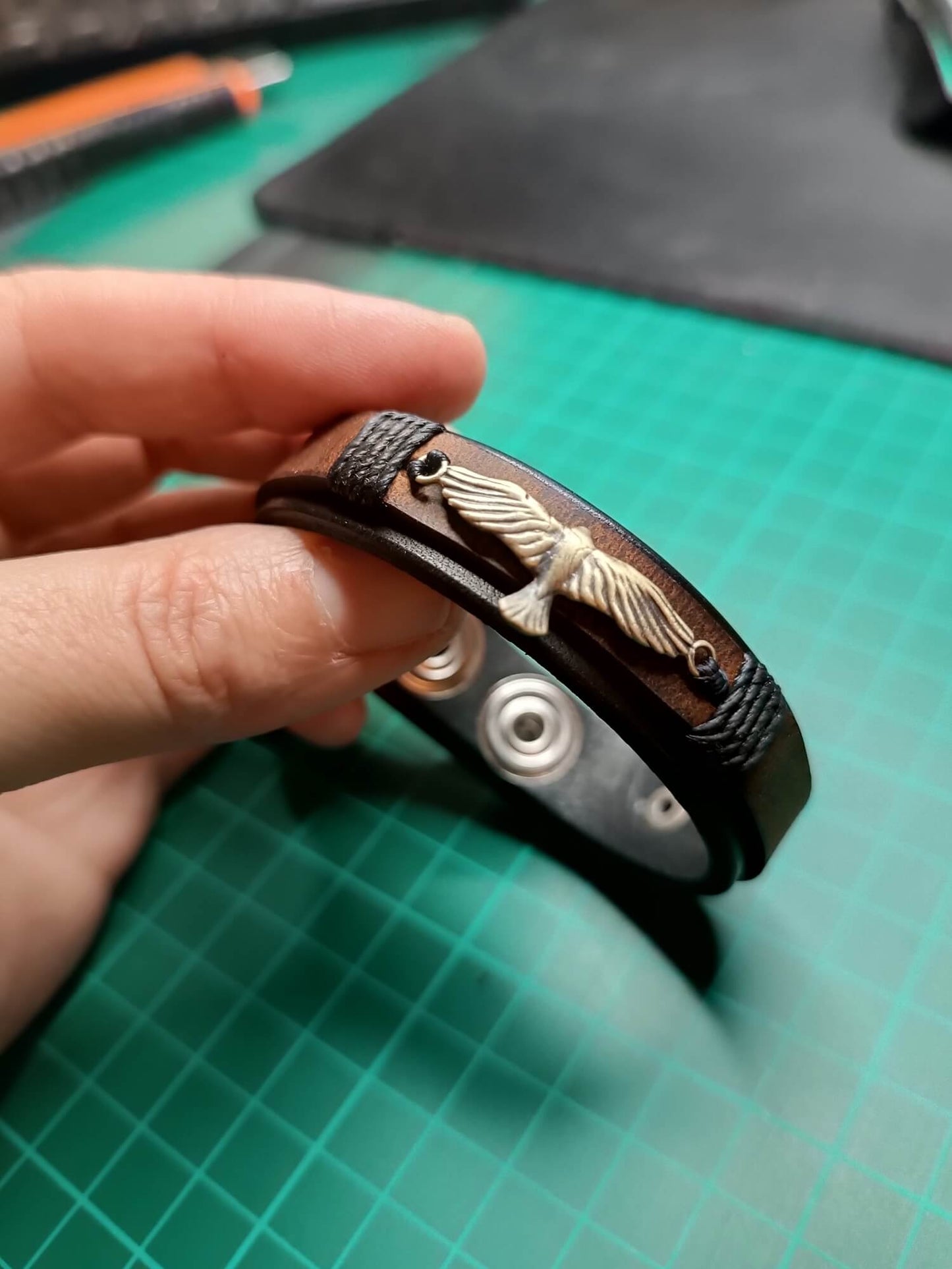 Hand holding a leather bracelet with eagle pendant on a green cutting mat.