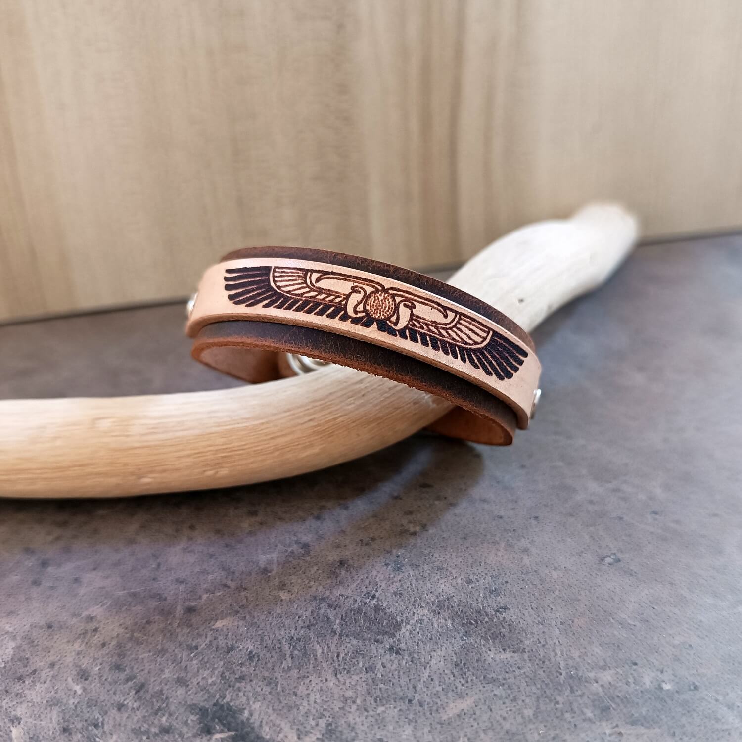Leather bracelet with egyptian winged disc design on a wooden stick against a neutral background