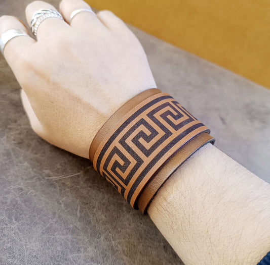 Hand wearing a brown leather cuff with Greek key design on a neutral background