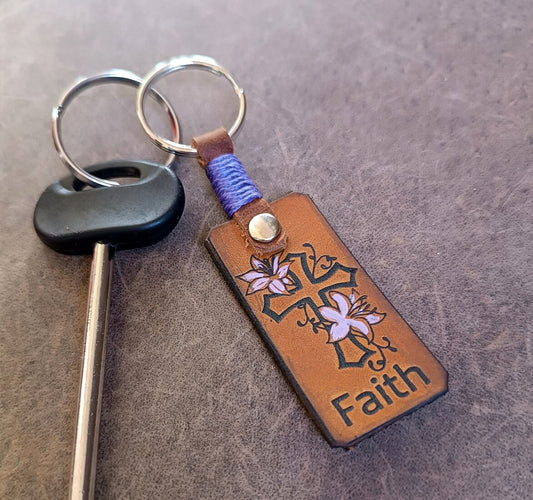 A brown leather keychain featuring a laser engraved and hand-painted floral cross with the word 'Faith' below it. The keychain also includes a personalized element, which appears to be a name in cursive script.