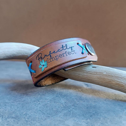 The Floral lioness Brown leather bracelet with 'Perfectly Imperfect' text close to the clasp and blue thread on a wooden surface.