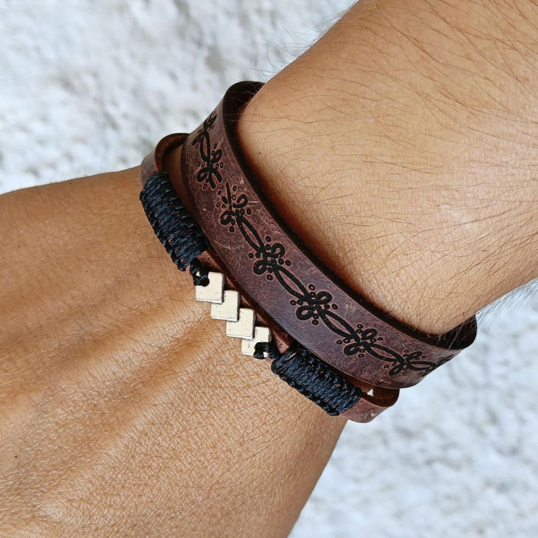 hand wearing a set of two leather bracelets, one dark brown with geometric laser engraved pattern and one thin with black macrame cord and geometric silver motif.
