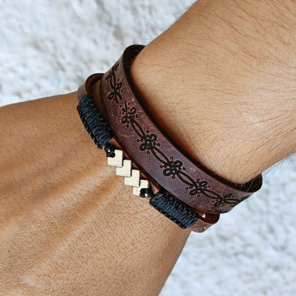 hand wearing a set of two leather bracelets, one dark brown with geometric laser engraved pattern and one thin with black macrame cord and geometric silver motif.
