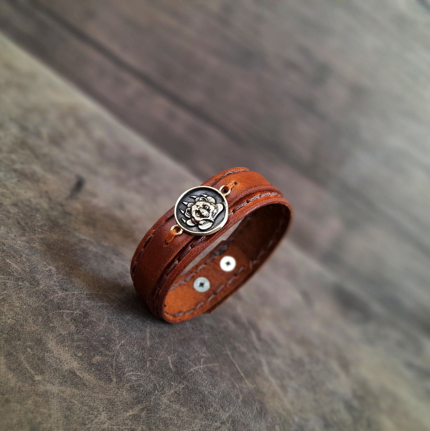 A brown leather bracelet with a layered style, featuring a grizzly bear design in the center, placed on a wooden surface.