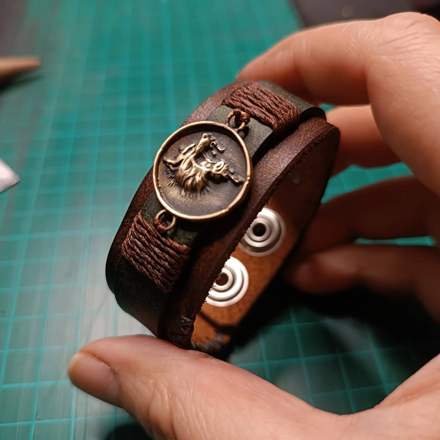 Brown leather bracelet with a growling wolf pendant held by a hand on a green cutting mat.