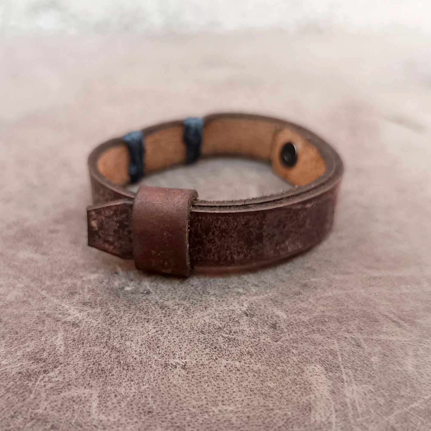 A handmade brown leather bracelet with a textured finish, gray stitched accents, and a metal snap closure, resting on a textured gray surface.
