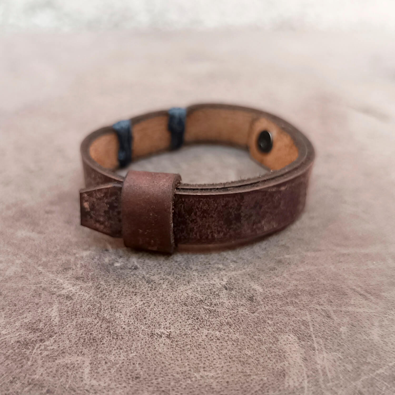 A handmade brown leather bracelet with a textured finish, gray stitched accents, and a metal snap closure, resting on a textured gray surface.