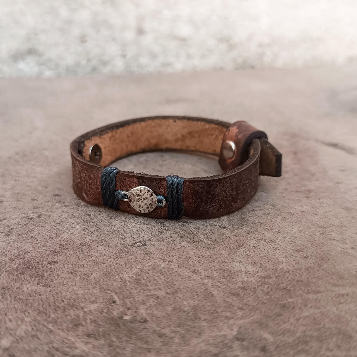 A handmade brown leather bracelet with dark gray cord wraps and a central round hammered moon  charm, resting on a textured gray surface.