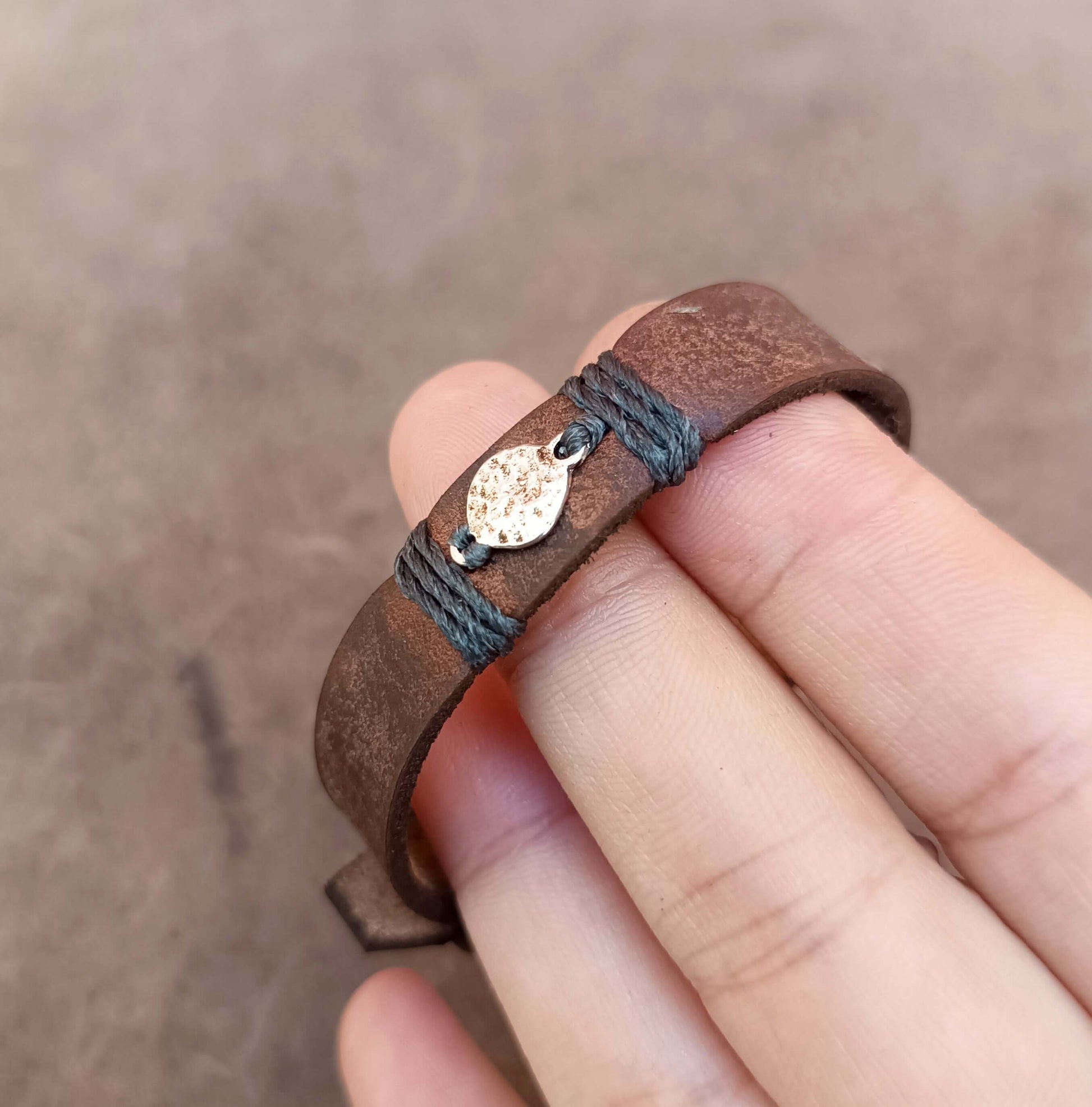 A close-up of a hand holding a brown leather wrap bracelet with dark gray cord wrapping and a small round hammered moon charm.