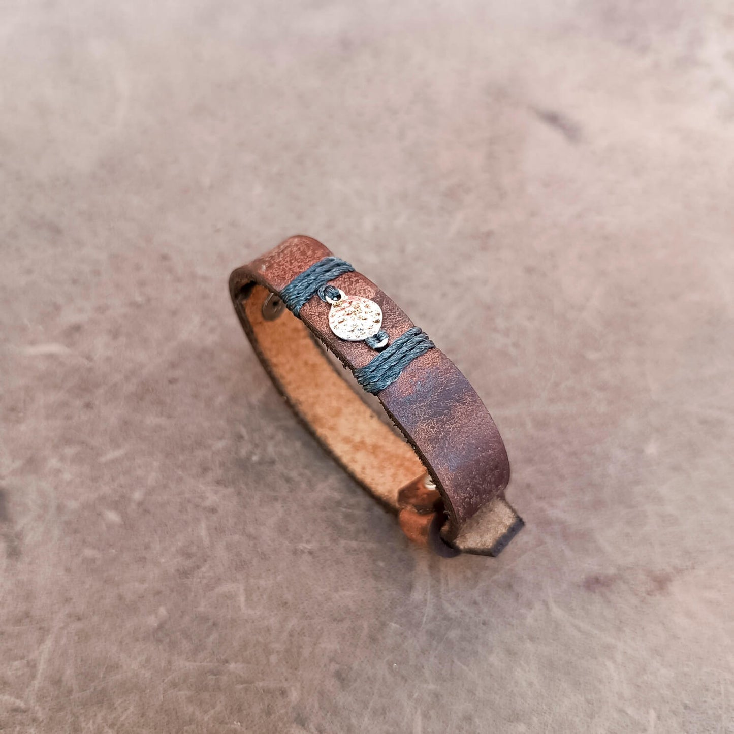 A handmade leather wrap bracelet with a distressed brown finish, wrapped with charcoal gray cord and featuring a small silver-toned hammered moon charm, resting on a textured gray surface.