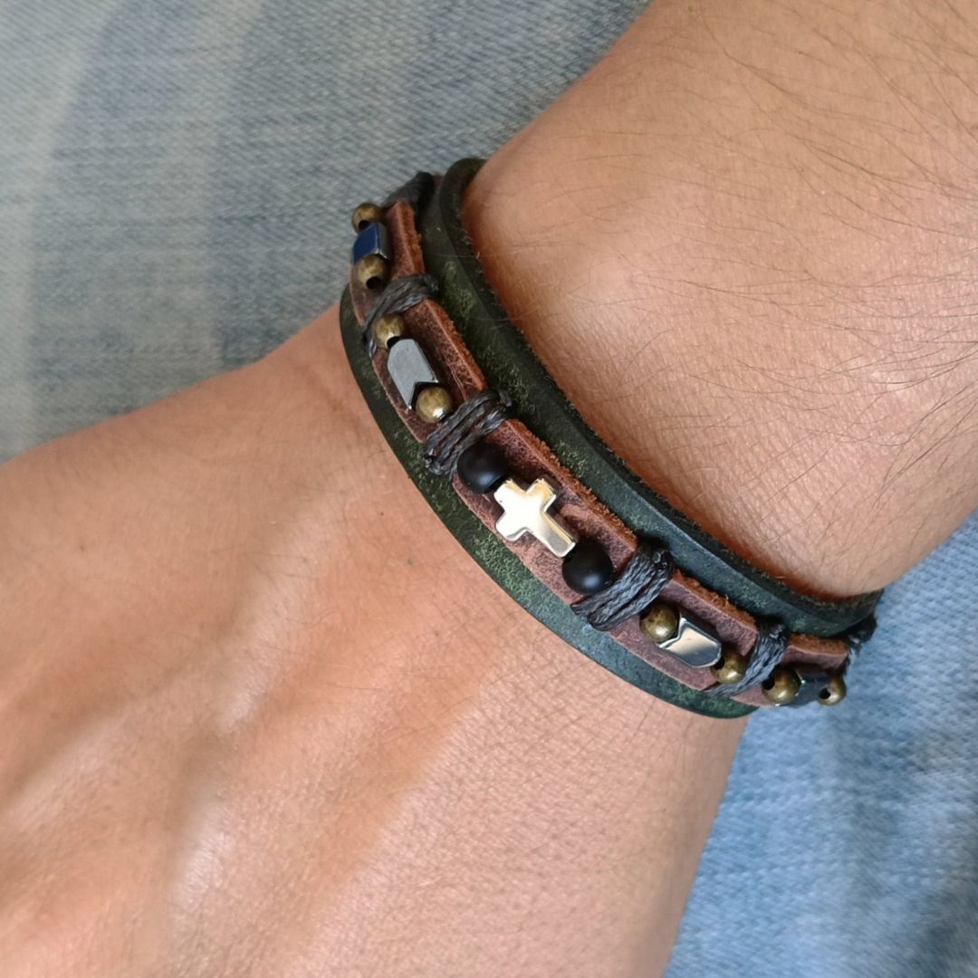 A close-up of a person's wrist wearing a multi-layered leather bracelet. The bracelet features dark brown leather, a dark green leather band, gray cord, beads, a silver-toned cross charm, set against a gray denim background.