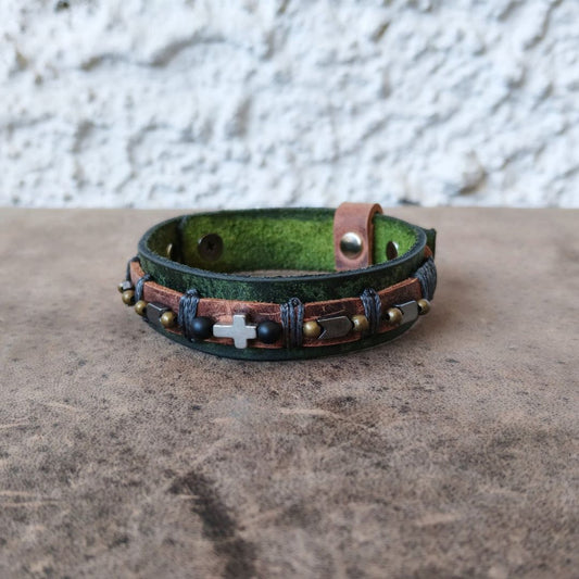 A handmade leather cuff bracelet with a distressed green and brown finish, decorated with black onyx beads, a silver hematite cross, and a silver-toned buckle, resting on a textured brown surface.