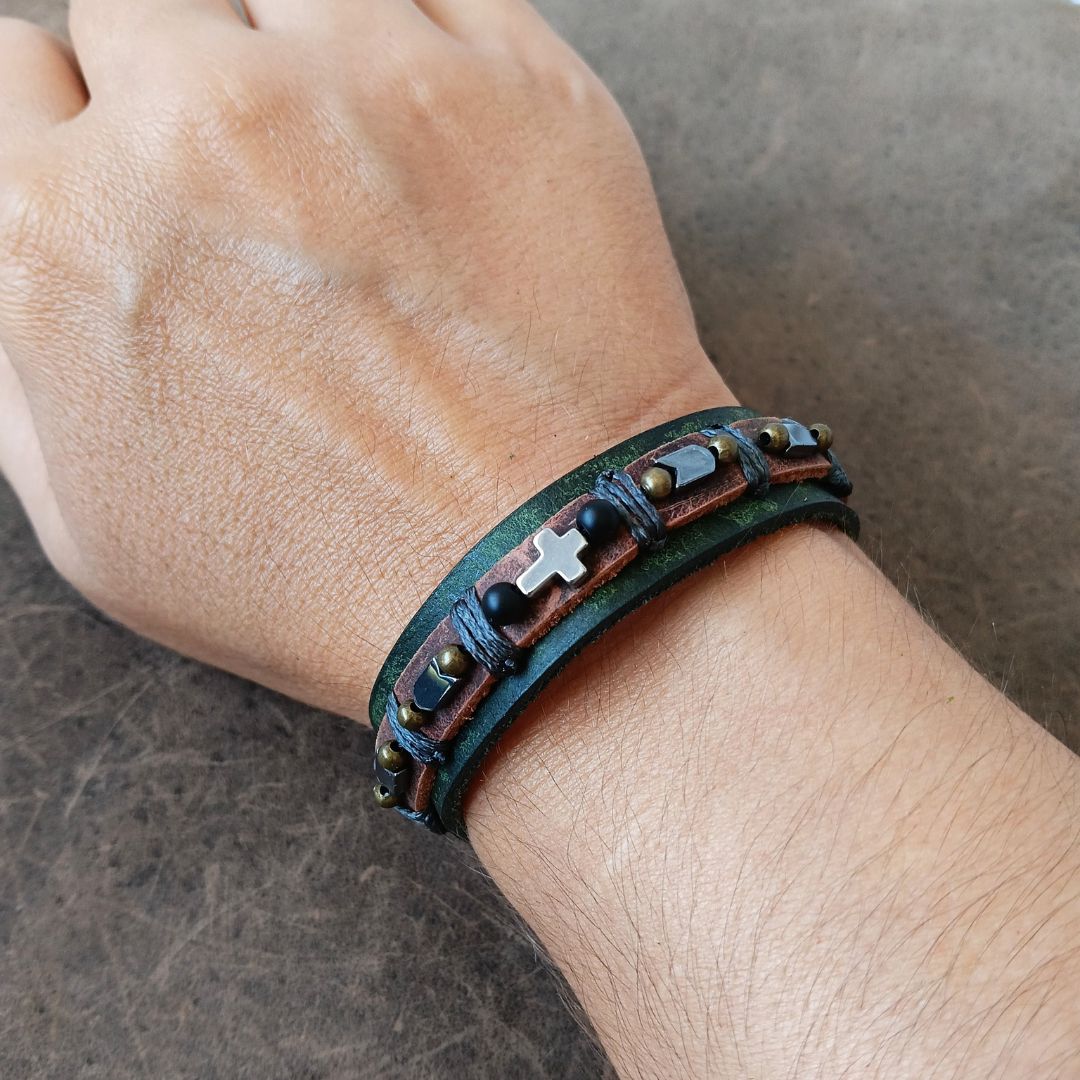 A close-up of a person's wrist wearing a dark green leather wrap bracelet with brown leather bands, small hematite beads, and a silver cross charm.