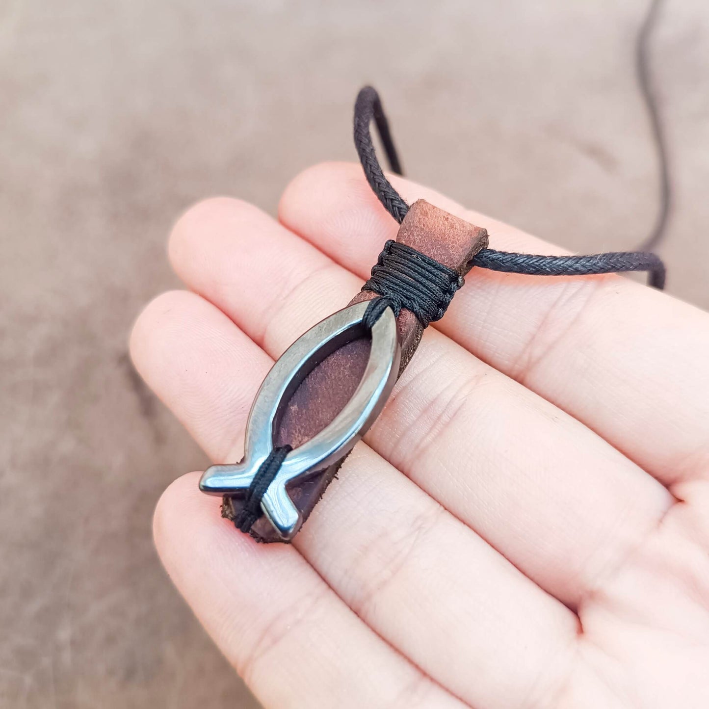 A small fish-shaped pendant necklace with hematite fish and dark brown leather wrap, held in a person's hand against a soft beige background.