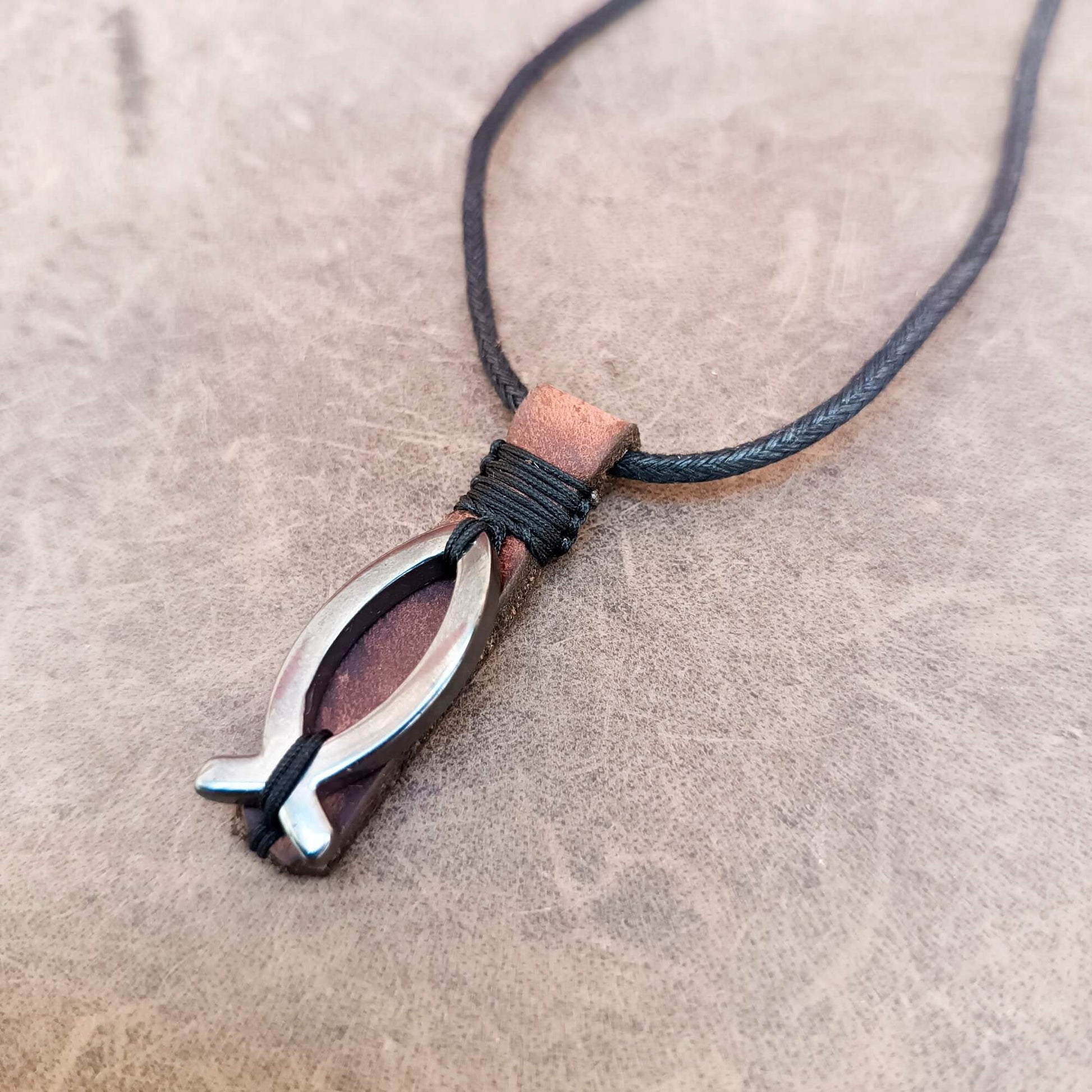 A close-up of a necklace with a dark brown leather cord and a pendant featuring a hematite fish-shaped charm over a reddish-brown leather backing, resting on a textured beige surface.