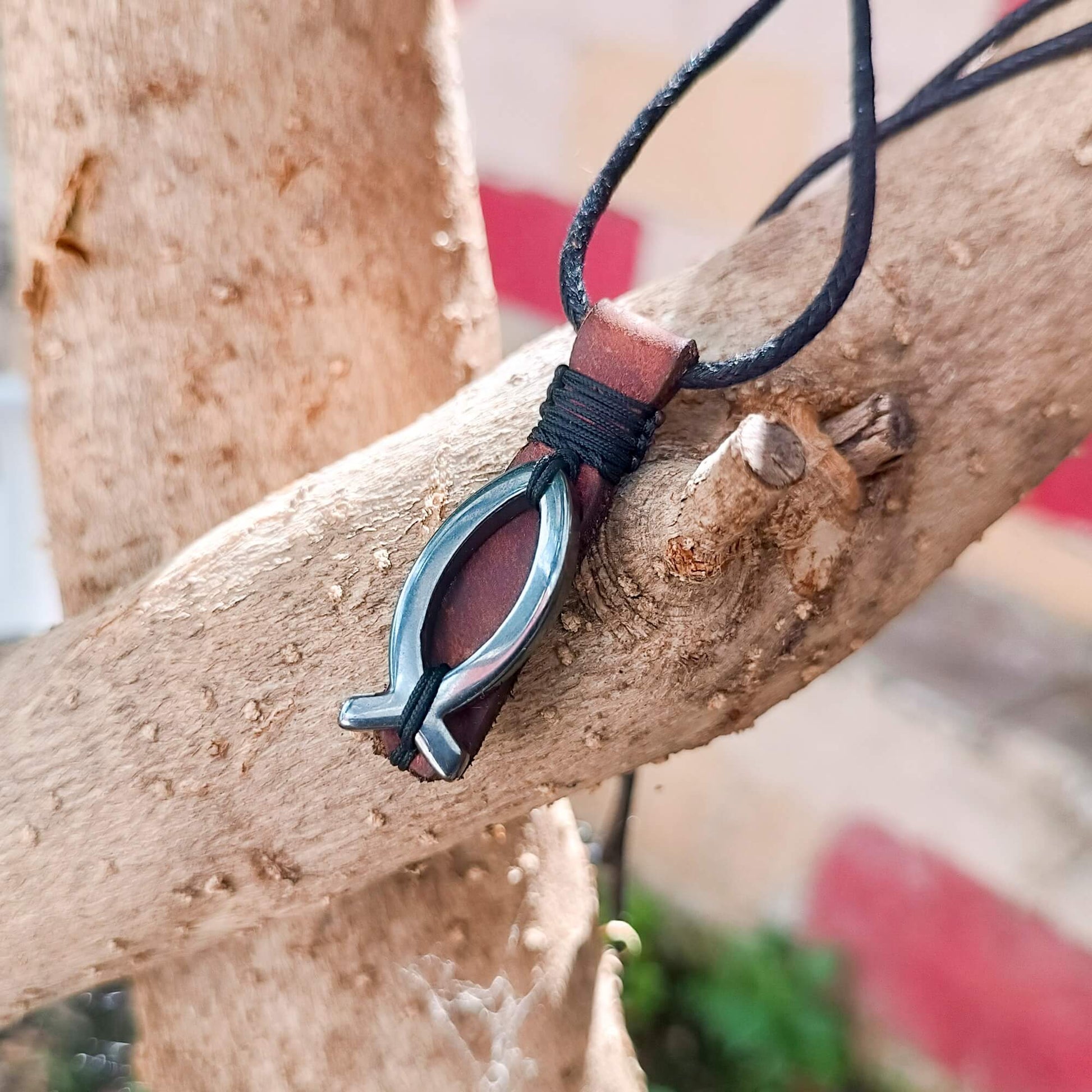 A close-up of a pendant necklace featuring a hematite fish wrapped in dark brown leather, hanging from a black cord and resting on a textured tree branch.