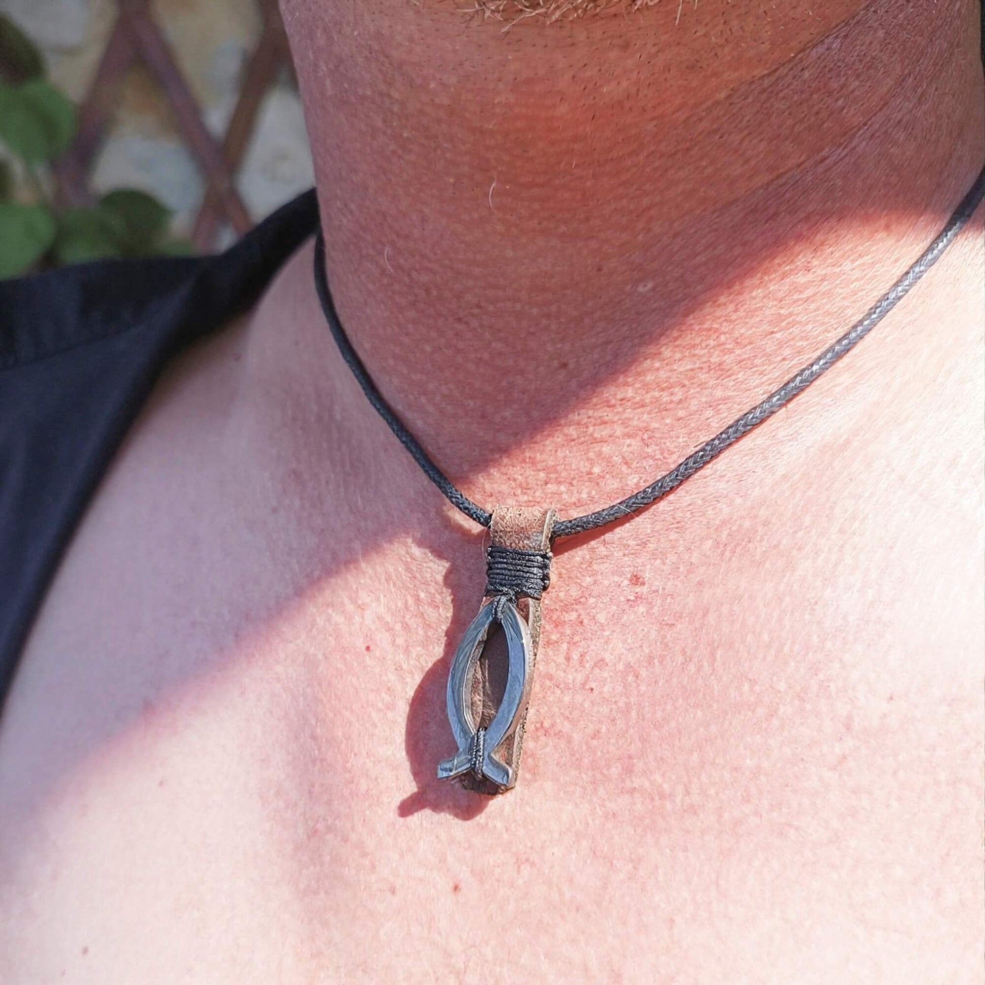 A close-up of a person's neck and chest wearing a dark cord necklace with a silver-toned hematite fish pendant.