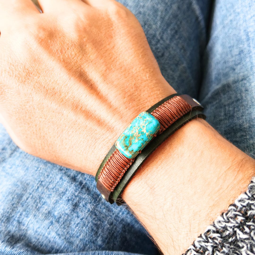 A close-up of a person's wrist wearing a handmade leather bracelet. The bracelet features a central turquoise-colored stone inlay with brown matrix, flanked by two brown wrapped leather bands and a black outer leather edge, set against a background of blue denim jeans.