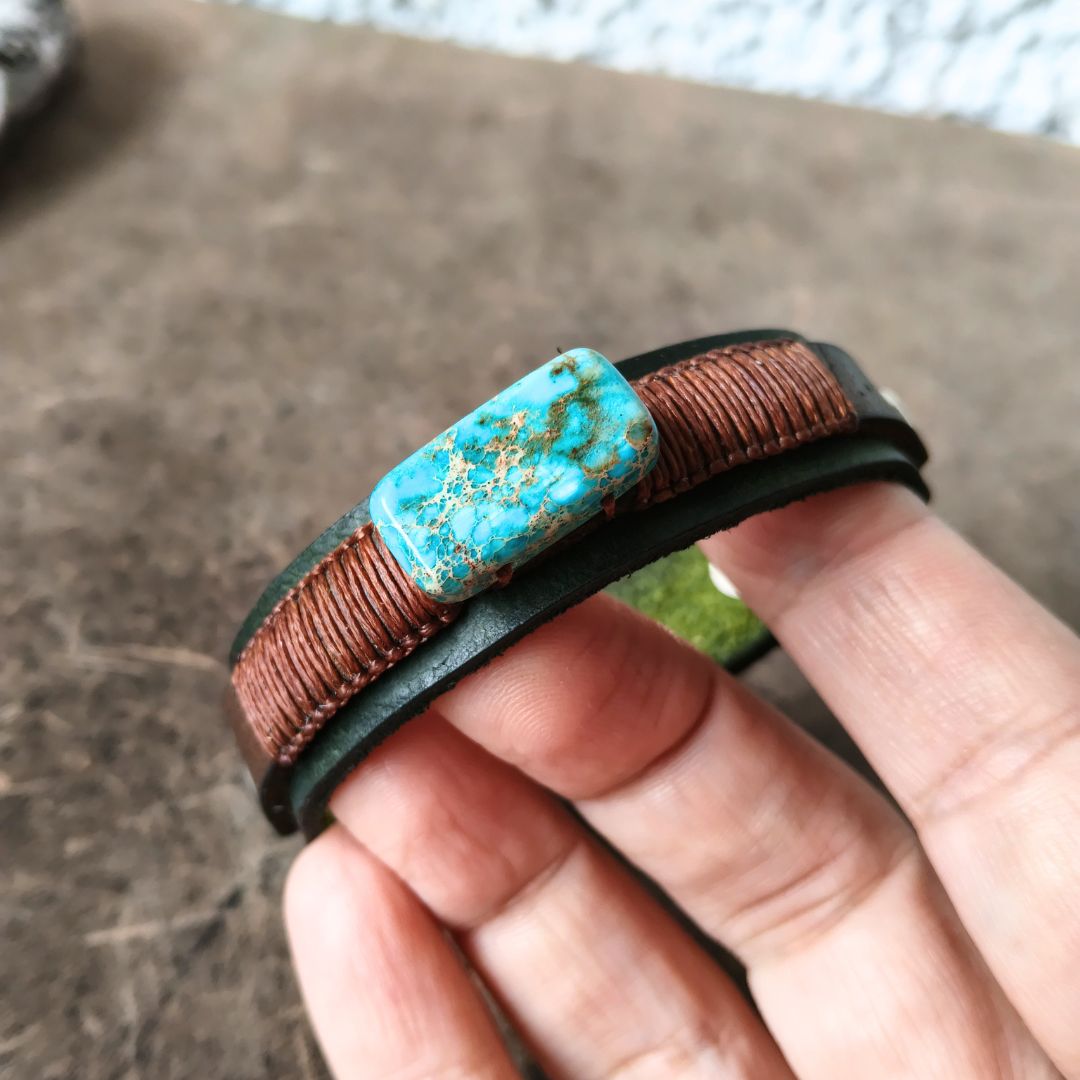 A close-up of a hand holding a brown leather wrap bracelet with a rectangular imperial jasper stone centerpiece and gold veining.