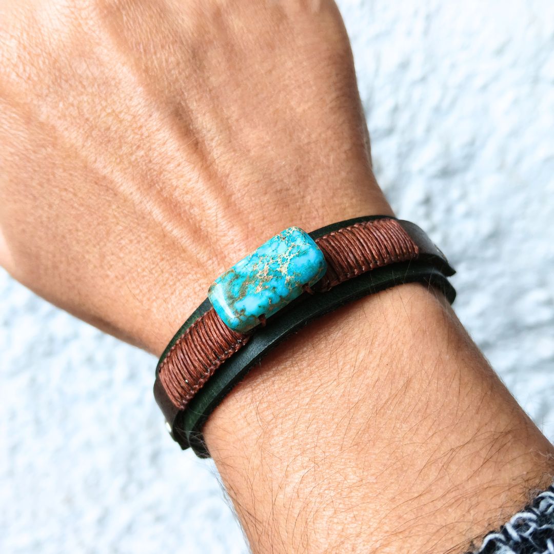 A close-up of a person's wrist wearing a multi-layered bracelet. The bracelet features a central rectangular imperial jasper stone with gold flecks, flanked by a brown wrapped cord and a dark green leather band, set against a light textured background.