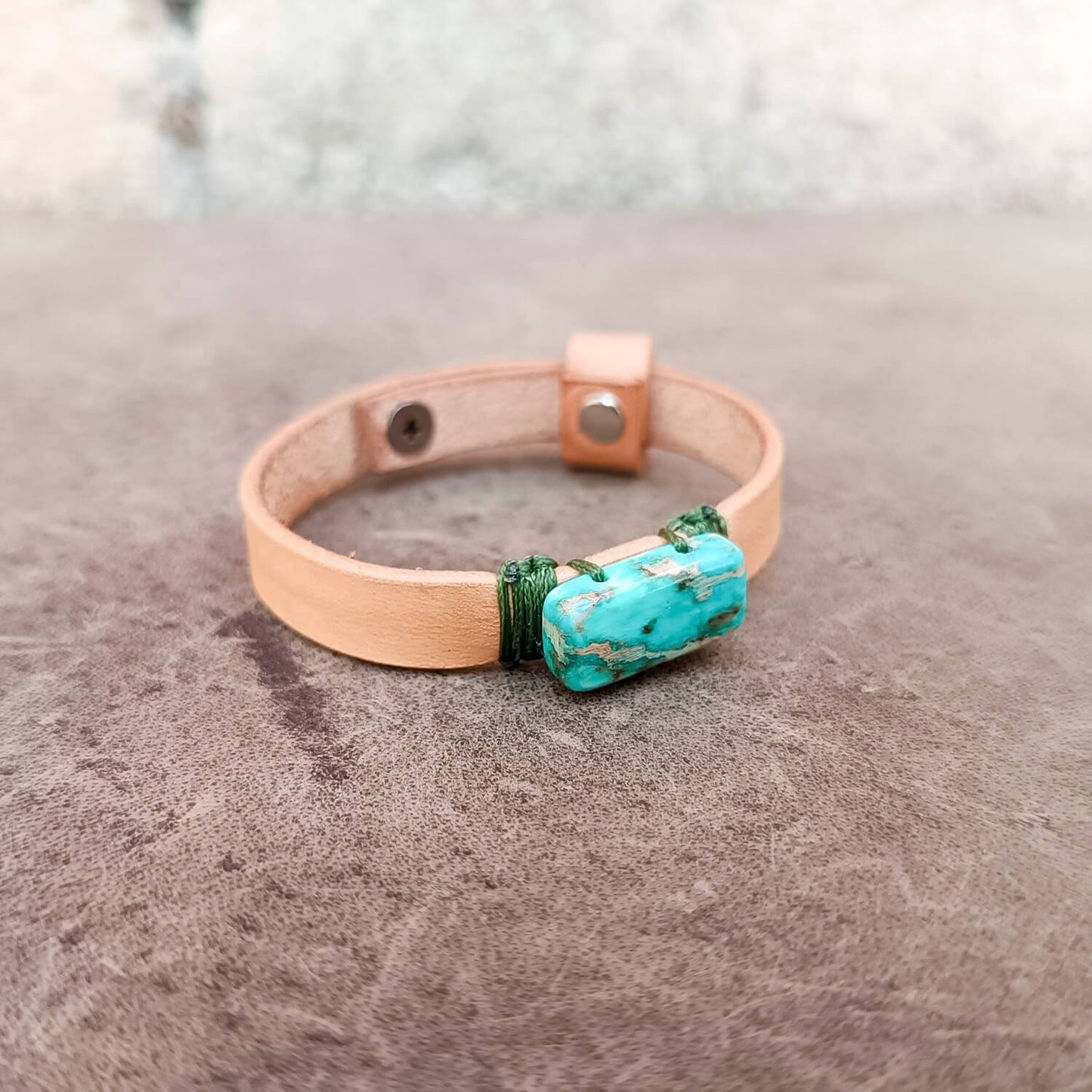 A handmade leather bracelet with Imperial Jasper stone centerpiece, wrapped in green cord, resting on a textured gray surface.