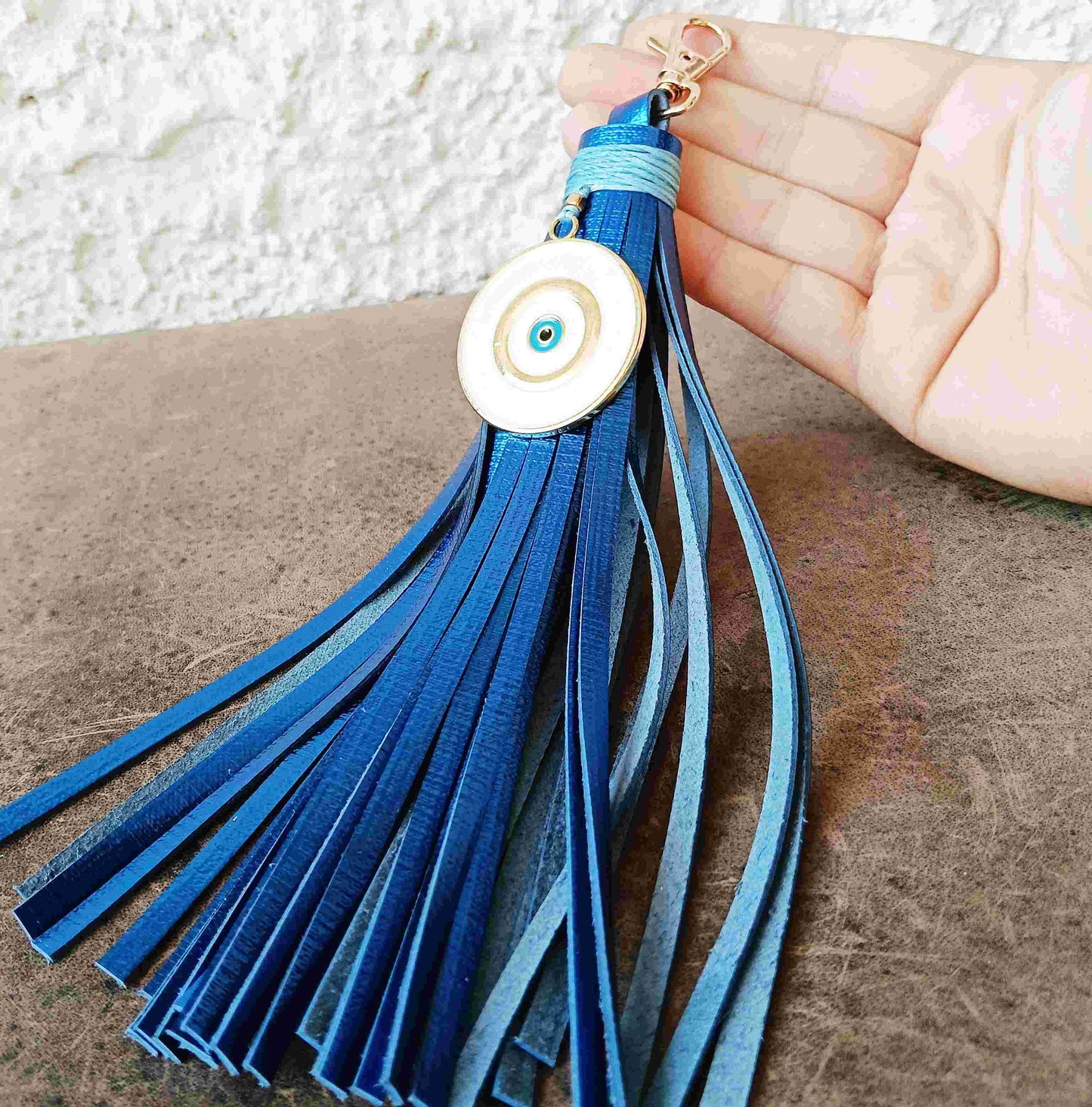 A blue leather tassel keychain with a circular white and blue evil eye charm, held in a hand over a brown textured surface.