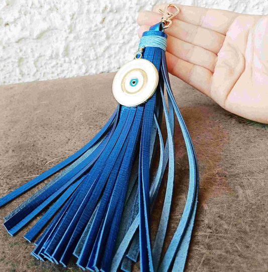 A blue leather tassel keychain with a circular white and blue evil eye charm, held in a hand over a brown textured surface.